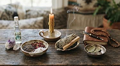 Traditional ritual items arranged on a wooden table, including a lit candle, sacred powders, herbs, and prayer beads